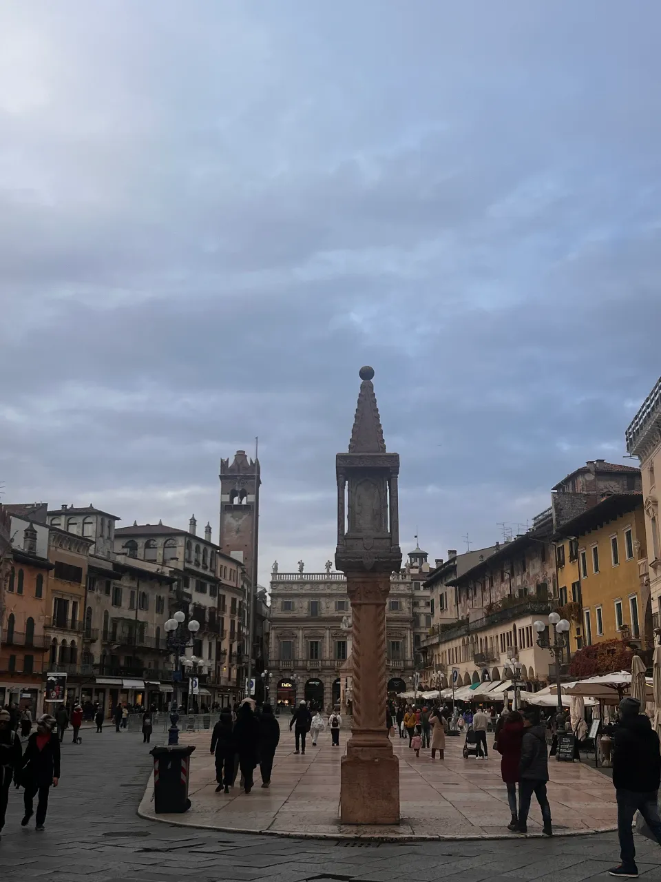 A stone monument, historic façades and pedestrians in Piazza delle Erbe under a cloudy winter sky