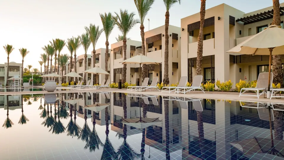 Sentido Reef Oasisの長いプール。淡い低層棟の前で、ヤシと白いパラソルが深青の水面に映っている