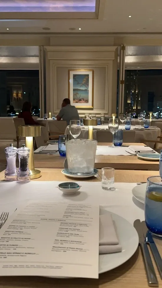 Rivieraの白いプレースマットと青いグラス、金色の卓上灯。奥に窓際の食事席が並ぶ