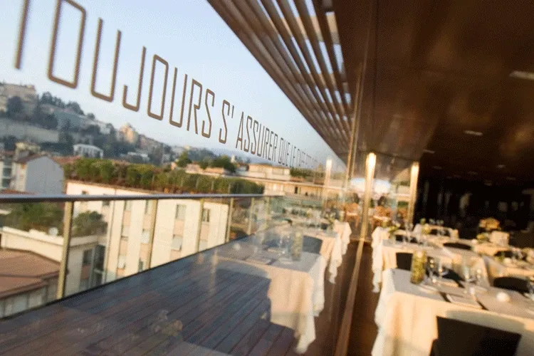 サン・マルコのRoof Garden Restaurant、ガラス張りの窓沿いの食卓と窓の向こうの丘の上のベルガモ
