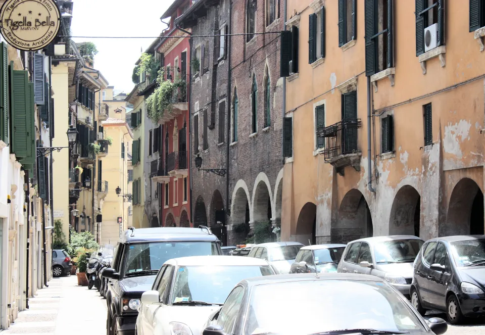 Low arches, brick façades and green shutters along Via Sottoriva in Verona, with parked cars in the foreground
