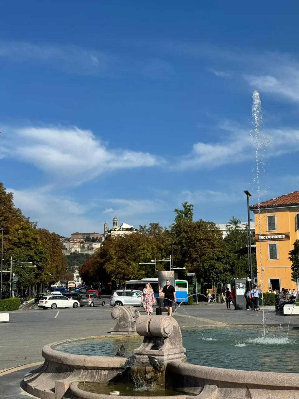 ベルガモ駅前の広場にある噴水と並木道、その奥に見える丘の上の旧市街