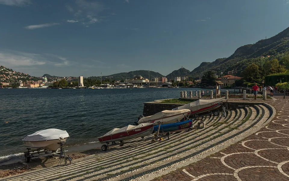 Pebbled lakeside steps, small boats and a promenade in Como, with the lake and wooded hills beyond