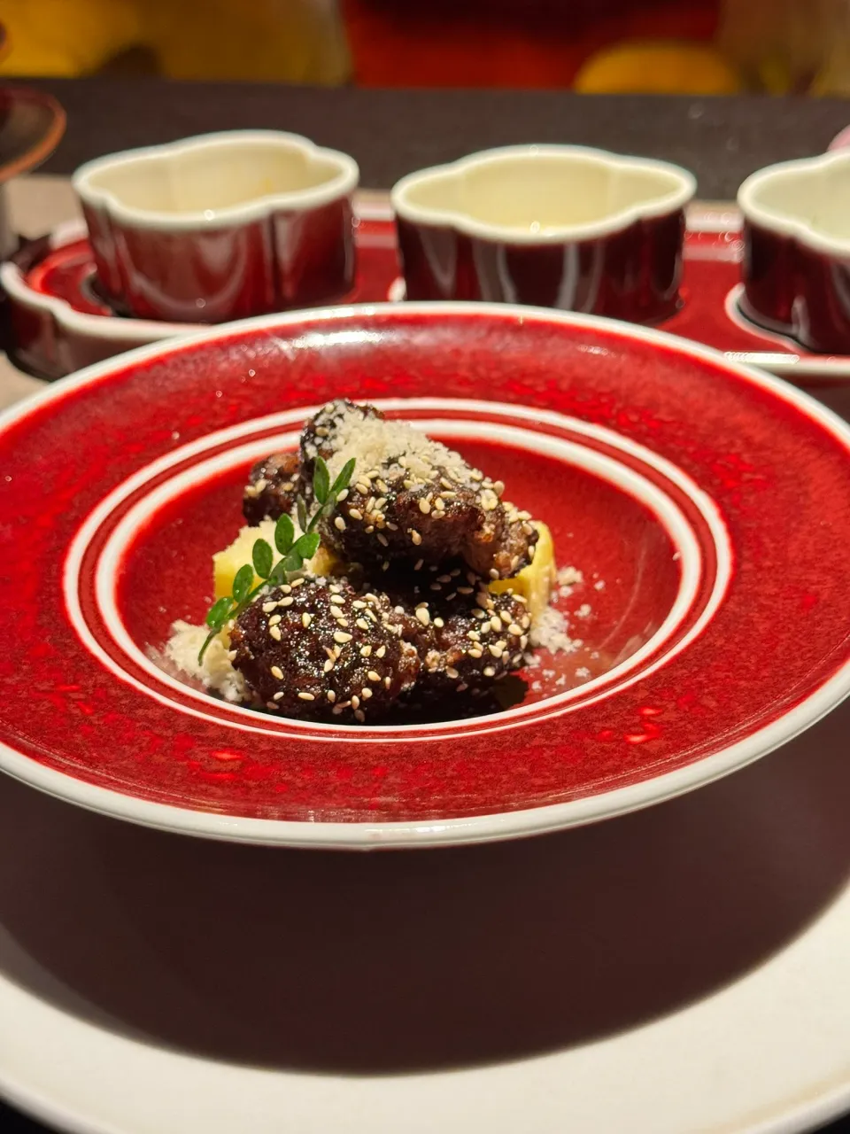 朱赤の器に盛られたごま付きの料理と奥に並ぶ三つの小鉢
