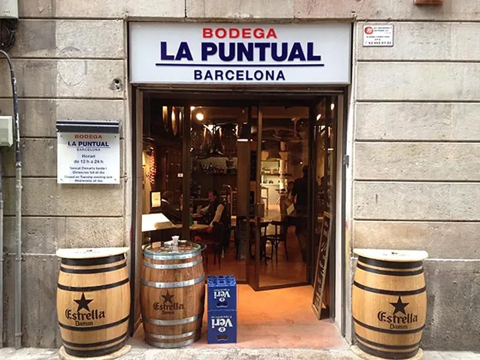 Bodega La Puntualの外観。赤と青の店名看板と、入口前の樽テーブル。
